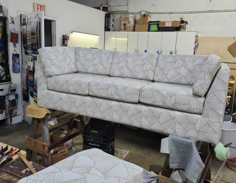 A patterned beige sofa is balanced on wooden sawhorses in a workshop, surrounded by tools, an ottoman with matching fabric, cabinets, and supplies. The workspace appears organized and busy.