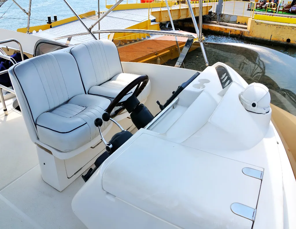 The helm of a white boat with two cushioned seats, a steering wheel, control levers, and navigational equipment, docked near a yellow pier on a sunny day.