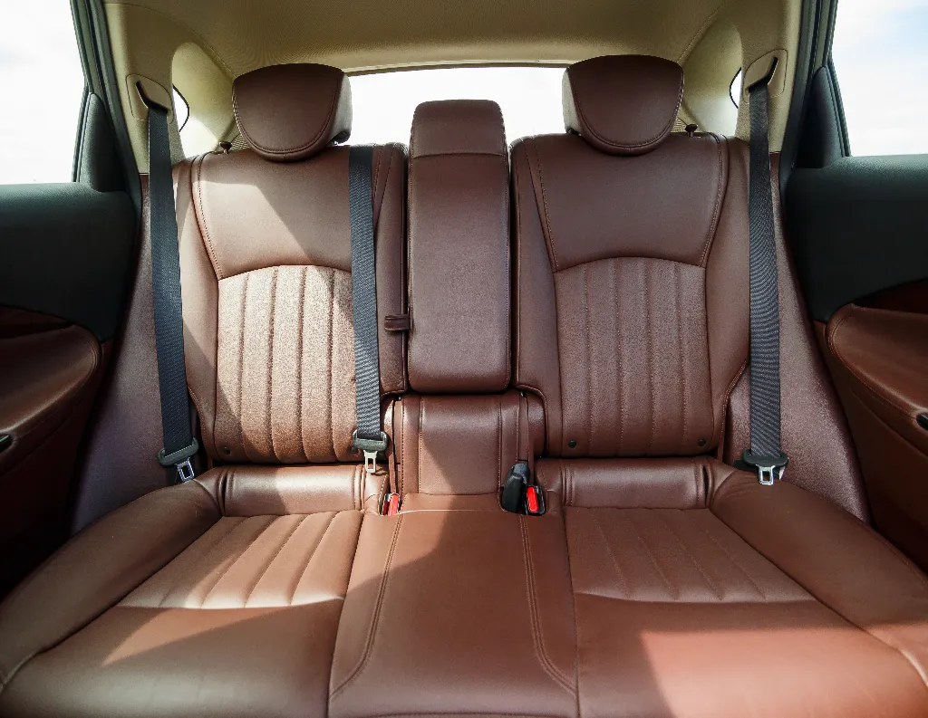 Brown leather backseat of a car with three seats, two seatbelts on the sides, a center armrest, and stitching details visible in the upholstery. Sunlight enters from the windows, highlighting the seats.