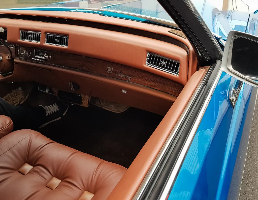The interior of a classic blue car with brown leather seats, a wood-trimmed dashboard, and vintage air vents. Part of a person’s leg wearing a black sneaker is visible in the passenger seat.