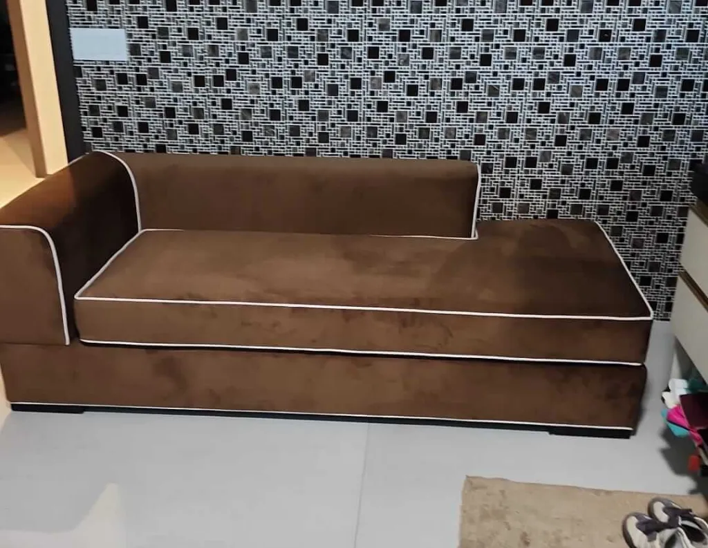 A brown L-shaped sofa with white piping sits in front of a black-and-white geometric patterned wall in a modern living room with a tiled floor.