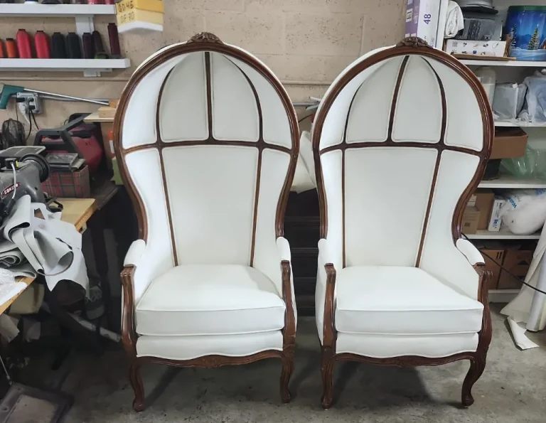 Two white, high-backed dome chairs with wooden frames sit side by side in a workshop, surrounded by tools, fabric, and upholstery supplies.