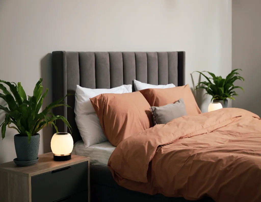 A neatly made bed with rust-colored bedding and multiple pillows, flanked by two nightstands with potted plants and glowing spherical lamps in a modern, minimalist bedroom.