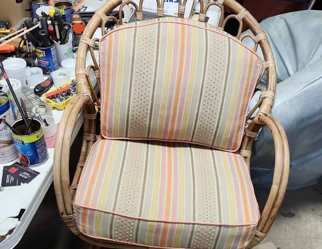 A wicker chair with a striped cushioned seat and backrest in orange, yellow, and beige tones sits next to a cluttered table with cans, bottles, and various items.