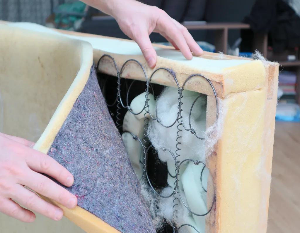 Close-up of hands pulling back the fabric and foam padding of a mattress, revealing metal springs and internal filling materials inside.