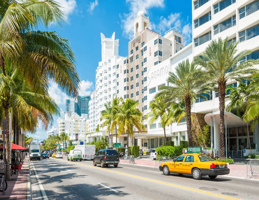 A sunny street in Miami Beach with palm trees, art deco buildings, cars, and a yellow taxi driving along the road under a bright blue sky.