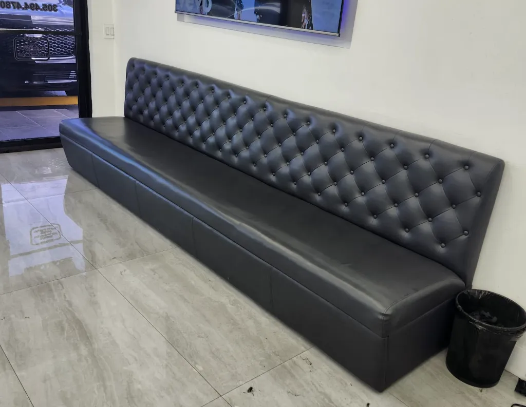 A long, dark gray, tufted bench sits against a white wall in a room with shiny tiled floors. A small black trash can is placed near one end of the bench. A door and a window are visible in the background.