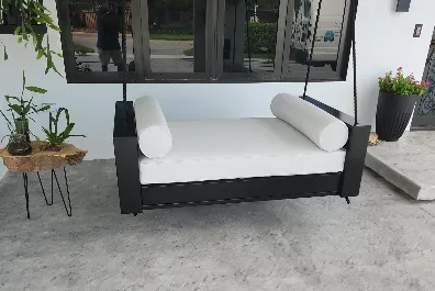 A modern outdoor swing bed with white cushions and bolsters, suspended by black ropes in front of a window, with plants on either side on a concrete patio.