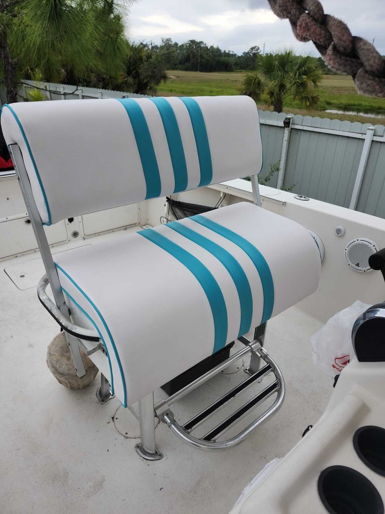 A white and turquoise cushioned boat seat with metal frame sits on a white boat deck, surrounded by a fence, palm trees, and greenery in the background. A large rock is placed behind one leg of the seat.