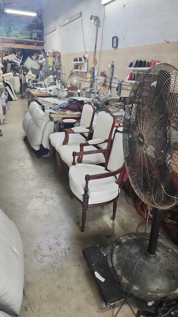 A workshop with white upholstered chairs, a large floor fan, various furniture, tools, and fabric rolls scattered around. Shelves and workbenches line the walls, creating a busy, cluttered workspace.