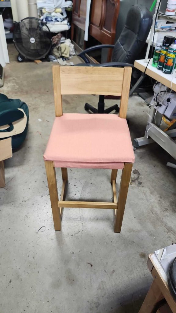 A wooden chair with a light pink cushioned seat is placed on a concrete floor in a cluttered workshop with scattered tools, shelves, a fan, and other furniture in the background.
