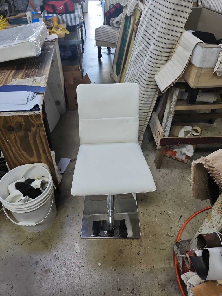 A white cushioned chair with a square chrome base sits on a concrete floor in a cluttered workshop, surrounded by fabric, tools, a white bucket, and various materials.