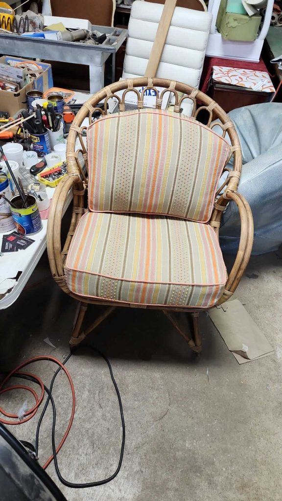 A wicker chair with curved arms and striped, multicolored cushions sits on a concrete floor in a cluttered workspace, surrounded by tools, cables, and miscellaneous items.