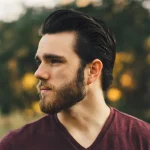 A man with dark, slicked-back hair and a full beard looks to his left. He is wearing a maroon V-neck shirt and is outdoors with blurred greenery and warm light in the background.