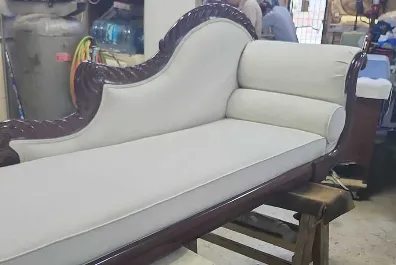 An elegant vintage chaise lounge with carved dark wood frame and light beige upholstered cushions, placed in a workshop setting with tools and supplies in the background.