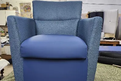 A blue upholstered armchair with wide armrests and a cushioned seat, placed indoors on a green carpet with various furniture and materials in the background.