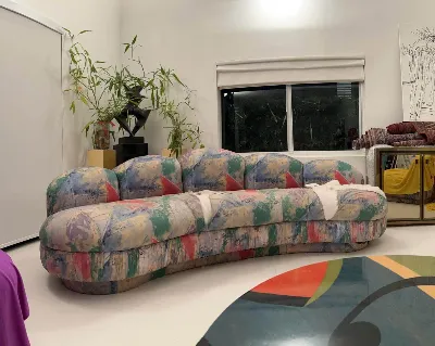 A pastel, abstract-patterned curved sofa sits in a modern living room with white walls, large window, potted plant, and colorful decor.