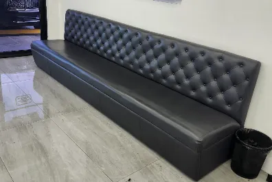A long, black, tufted leather bench with a high backrest sits against a white wall on a shiny tiled floor. A small black trash can is placed at one end of the bench.