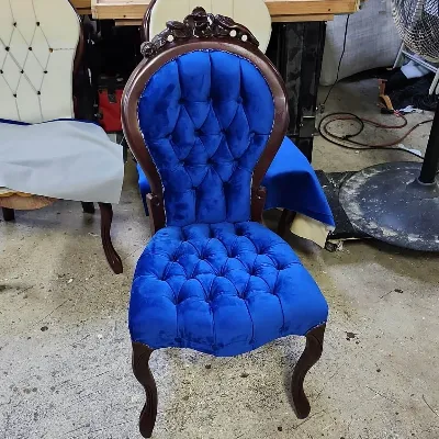 A vintage wooden chair with ornate detailing and blue tufted velvet upholstery sits on a workshop floor, with tools and materials visible in the background.