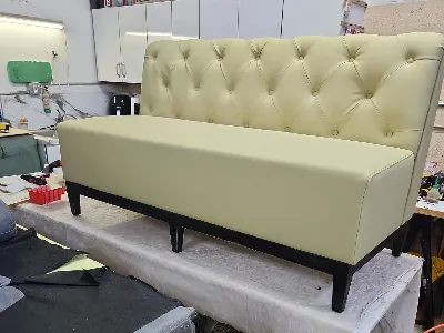 A light beige upholstered bench with a tufted backrest and black wooden legs is placed on a worktable in a workshop setting.
